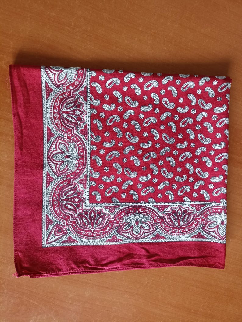 May include: A folded red bandana with a white paisley pattern. The bandana has a detailed border design with floral and scroll elements. The paisley pattern is repeated across the main field of the bandana.