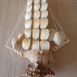 Op de afbeelding: Een decoratieve zeeschelp zeilboot met crèmekleurige zeilen en een grote, getextureerde schelpbasis. De zeilen zijn gemaakt van meerdere zeeschelpen en de boot is gemonteerd op een basis van kleine kiezels en schelpen.