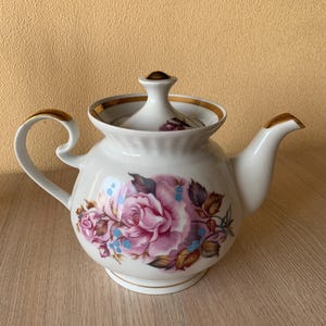 Könnte beinhalten: Eine weiße Porzellan-Teekanne mit goldener Verzierung und einem Blumendesign mit rosa Rosen und blauen Blumen. Die Teekanne hat einen Deckel mit einem goldenen Knopf.