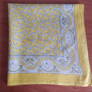 May include: Yellow paisley patterned handkerchief with a white and gray decorative border. The handkerchief is folded and lies on a wooden surface. The paisley pattern is repeated across the fabric.