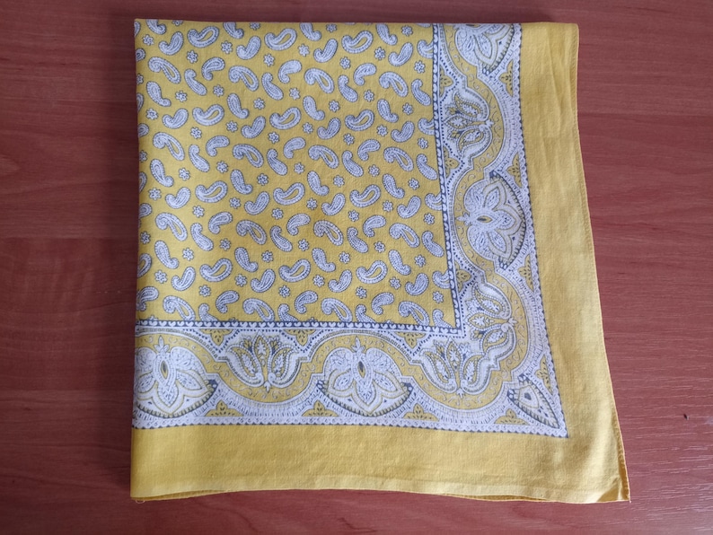 May include: Yellow paisley patterned handkerchief with a white and gray decorative border. The handkerchief is folded and lies on a wooden surface. The paisley pattern is repeated across the fabric.