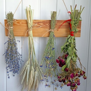 May include: A wooden hanging rack displays four bundles of dried flowers and grains. The bundles include lavender, wheat, blue cornflowers, and red roses. Each bundle is tied with twine or ribbon, creating a rustic, natural aesthetic.