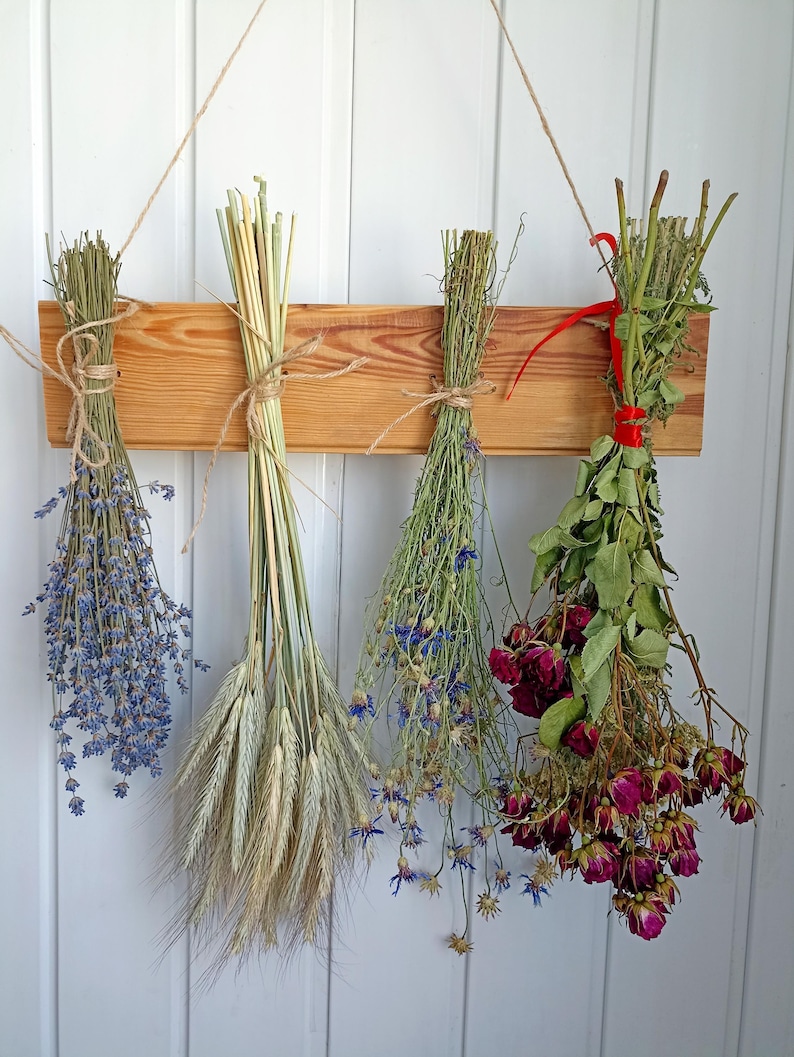 May include: A wooden hanging rack displays four bundles of dried flowers and grains. The bundles include lavender, wheat, blue cornflowers, and red roses. Each bundle is tied with twine or ribbon, creating a rustic, natural aesthetic.