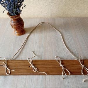 May include: A wooden wall hanging with three jute twine ties. The wood is a warm brown color. A decorative brown vase with lavender flowers sits to the left of the wall hanging. The vase has carved details.