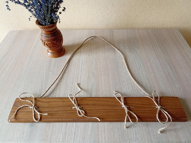 May include: A wooden wall hanging with three jute twine ties. The wood is a warm brown color. A decorative brown vase with lavender flowers sits to the left of the wall hanging. The vase has carved details.