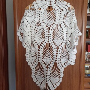 May include: A white, open-weave crochet shawl with a scalloped edge, displayed on a wooden hanger. The intricate lace-like pattern features geometric and floral motifs, creating a delicate and elegant design.