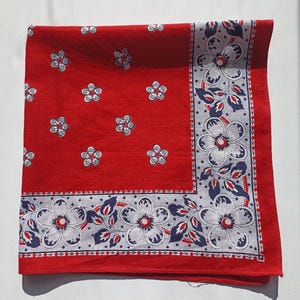 May include: A red square bandana with a white and blue floral border and scattered white flowers. The bandana is folded, displaying the detailed floral pattern. The design includes white flowers with blue accents.