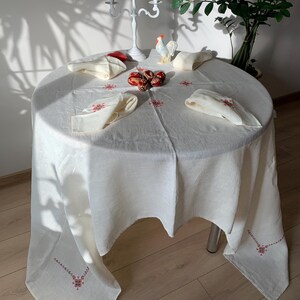 May include: A white linen tablecloth with red embroidered floral designs. The tablecloth is laid on a round table with four place settings, each with a folded napkin. There are red Easter eggs and a white ceramic rooster on the table.