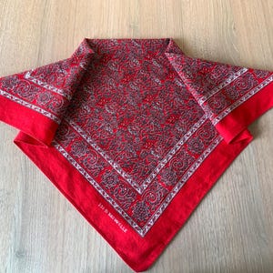 May include: Red bandana with a paisley pattern in shades of gray and white. The bandana has a white border with a decorative design. The text "100% Baumwolle" is printed on the edge of the bandana.