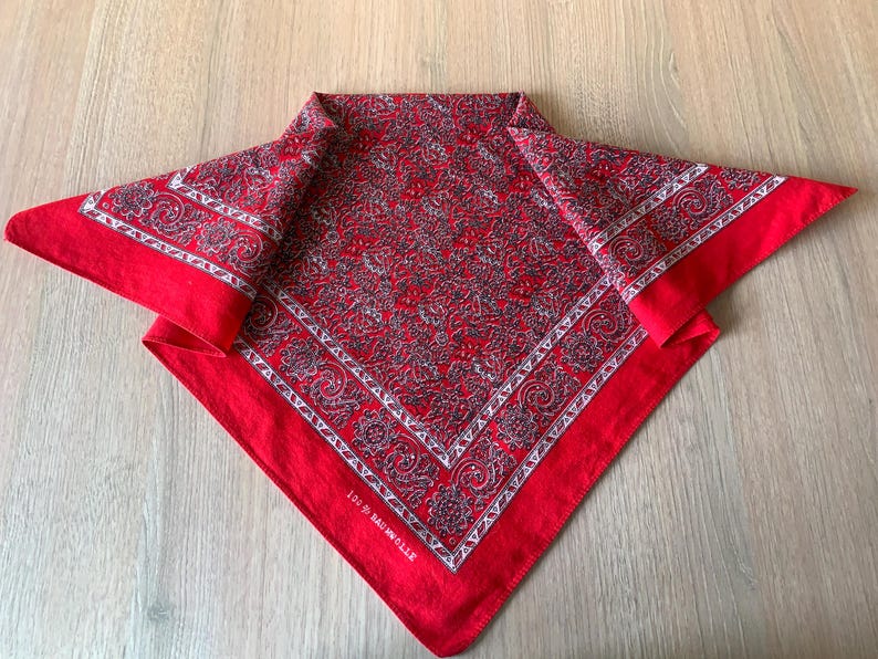 May include: Red bandana with a paisley pattern in shades of gray and white. The bandana has a white border with a decorative design. The text "100% Baumwolle" is printed on the edge of the bandana.