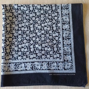 May include: A square, navy blue bandana with a white floral pattern covering most of the fabric. A border of white floral designs and a solid navy blue edge frame the pattern. The text "100% BAUMWOLLE/COTTON/COTON" is printed on the edge.