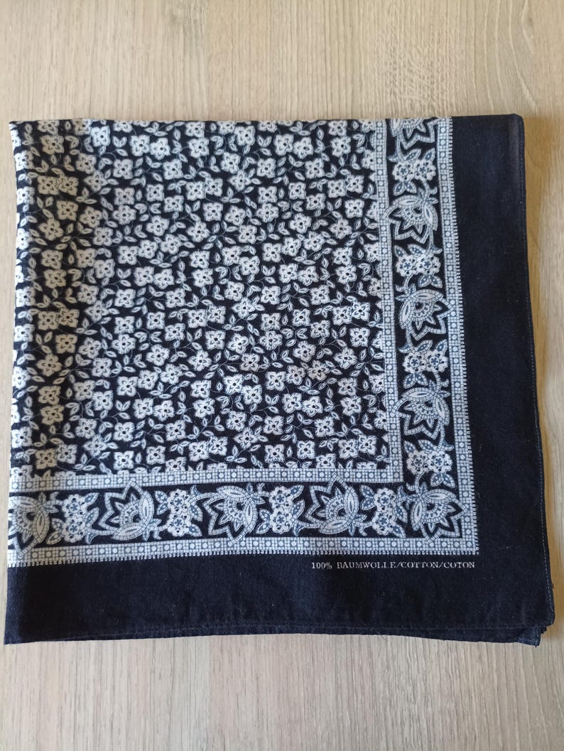 May include: A square, navy blue bandana with a white floral pattern covering most of the fabric. A border of white floral designs and a solid navy blue edge frame the pattern. The text "100% BAUMWOLLE/COTTON/COTON" is printed on the edge.