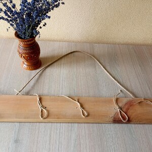 May include: A wooden wall-mounted coat rack with three jute rope loops for hanging items. The rack is made of light-colored wood and has a natural finish. A brown vase with lavender is in the background.