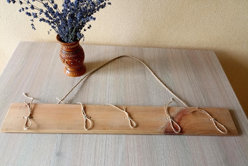 May include: A wooden wall-mounted coat rack with three jute rope loops for hanging items. The rack is made of light-colored wood and has a natural finish. A brown vase with lavender is in the background.