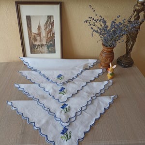 Può includere: Quattro fazzoletti bianchi con bordi smerlati blu e fiori blu ricamati. I fazzoletti sono disposti su una superficie di legno. Un'immagine incorniciata, un vaso di lavanda e un portacandele decorativo sono sullo sfondo.