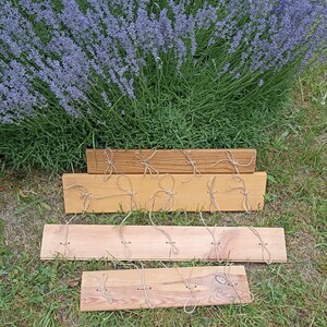 May include: Four wooden planks of varying shades of brown, each with twine loops attached. The planks are arranged on green grass, with a backdrop of a large lavender bush with purple flowers.