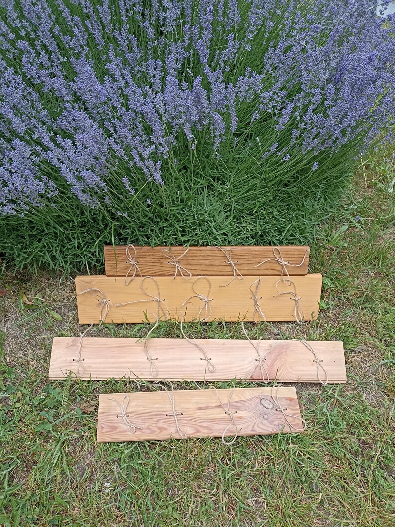 May include: Four wooden planks of varying shades of brown, each with twine loops attached. The planks are arranged on green grass, with a backdrop of a large lavender bush with purple flowers.