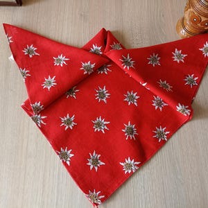 Puede incluir: Un pañuelo rojo con un estampado de flores de edelweiss blancas y amarillas. El pañuelo está doblado en forma triangular, mostrando el diseño floral. La tela parece ser de algodón o un material similar.