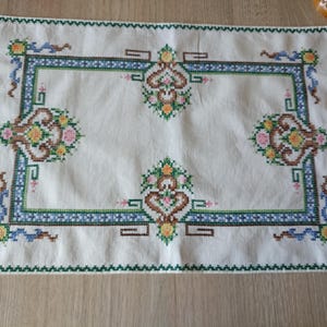 May include: A rectangular, off-white embroidered placemat with a detailed floral and geometric design. The border and central frame are green and blue, with floral motifs in pink, yellow, and brown. The edges are finished with a green zigzag stitch.