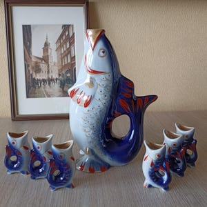 May include: A set of blue and white porcelain fish-shaped shot glasses and a matching decanter. The decanter is larger and has a red and gold design. The shot glasses are smaller and have a blue and white design.