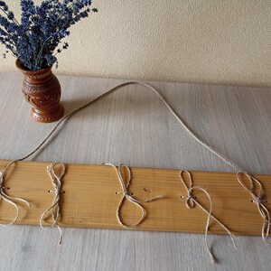 May include: A wooden wall hanging with five jute loops for hanging items. The wood is light brown, and a jute rope is attached for hanging. A small brown vase with dried lavender sits to the left.