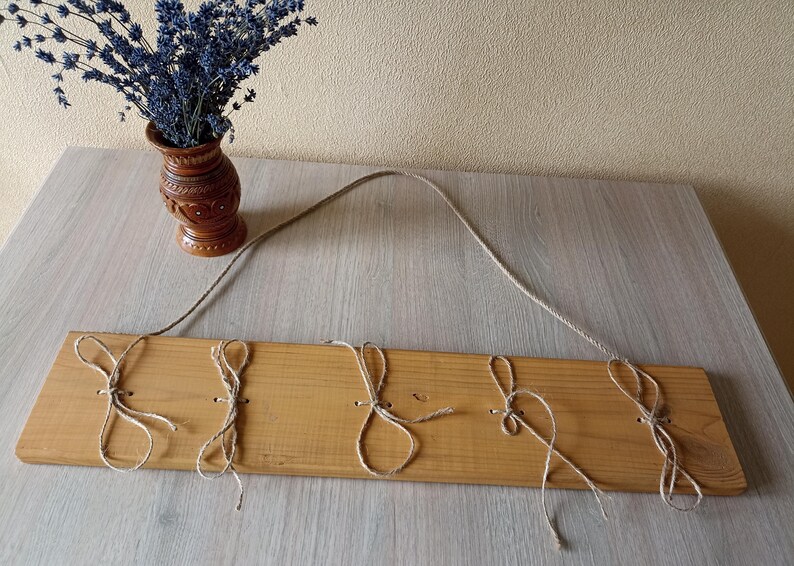 May include: A wooden wall hanging with five jute loops for hanging items. The wood is light brown, and a jute rope is attached for hanging. A small brown vase with dried lavender sits to the left.