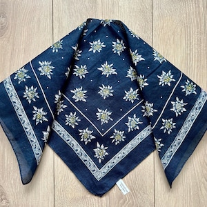 Può includere: Un bandana blu navy con un motivo floreale bianco e un bordo bianco. Il bandana è piegato a forma quadrata ed è realizzato in un tessuto leggero. Il motivo floreale è distribuito uniformemente sul tessuto.
