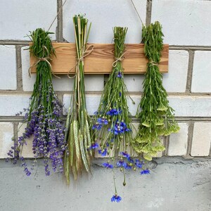 May include: A wooden wall hanging featuring four bundles of dried flowers and herbs. The bundles include lavender, wheat, blue cornflowers, and green herbs, tied with twine. The wooden plank is suspended by twine against a white brick wall.