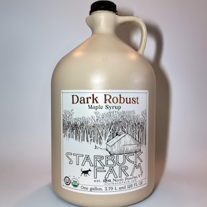 Puede incluir: Una jarra beige de un galón (3,79 L) de jarabe de arce Dark Robust de Starbuck Farm. La etiqueta presenta una escena de bosque y una ilustración de granero. La jarra tiene una tapa negra y un asa.