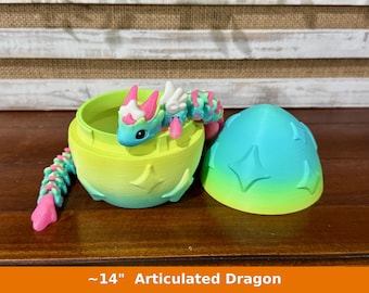 May include: An articulated dragon with a blue, pink, and white body emerging from a two-piece egg container. The egg is colored in a gradient of lime green to turquoise blue and features embossed star shapes on the exterior surface.