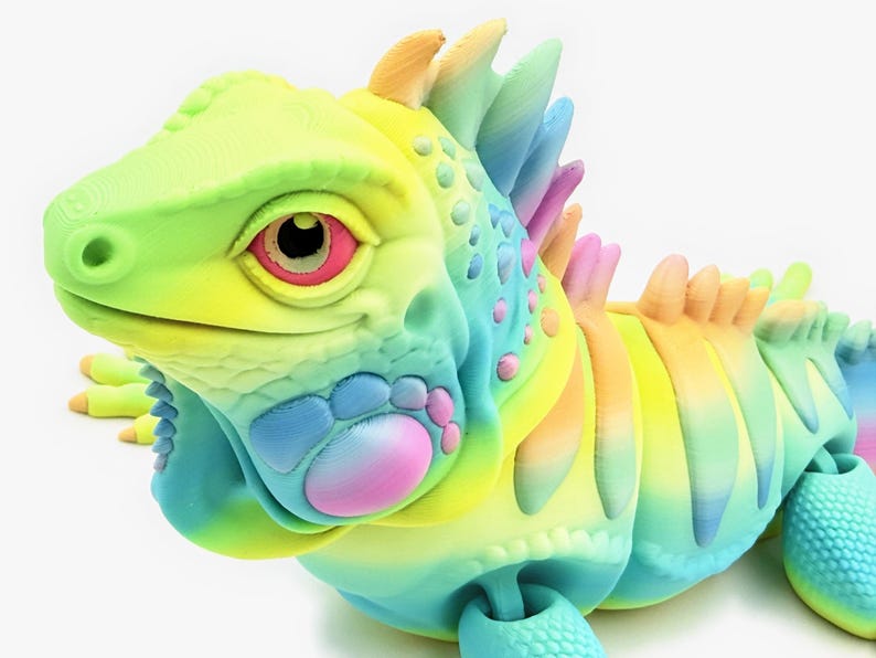 May include: A 3D printed articulated iguana figurine featuring a vibrant rainbow gradient color scheme. The lizard has a textured surface with prominent scales, a spiky crest along its back, and large eyes with pink and black details.