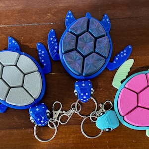 May include: Three blue and green turtle-shaped keychains with different colored shells. The shells are segmented and have a textured surface. The keychains are attached to silver key rings.