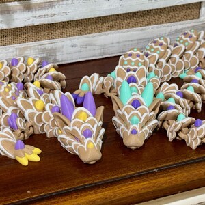 May include: Two gingerbread dragon figurines with intricate details. One dragon is decorated with purple and yellow accents, while the other is decorated with turquoise and purple accents.