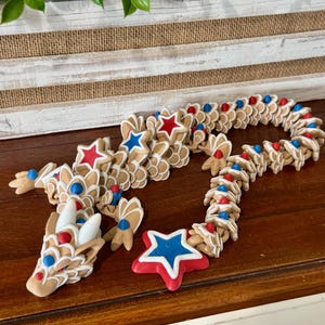 May include: A decorative dragon-shaped garland with a patriotic theme. The garland features star-shaped and petal-shaped elements in shades of tan, red, white, and blue. The dragon's head and tail are also visible, with a star-shaped element at the end.