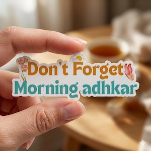 May include: A white sticker with the text "Don't Forget Morning adhkar" in orange and teal. The sticker features floral and butterfly illustrations. The sticker is held by a hand.