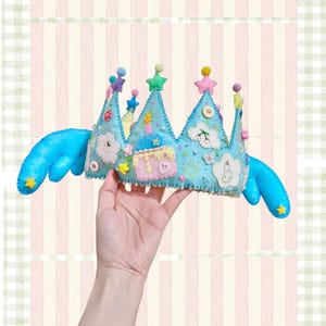 Puede incluir: Una corona caprichosa de fieltro azul claro con adornos de estrellas y nubes, un pastel rosa y alas azules. La corona está adornada con cuentas y estrellas de colores. Una mano sostiene la corona sobre un fondo a rayas.