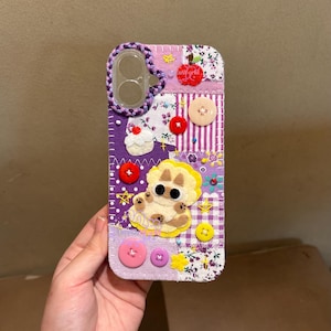 May include: A patchwork phone case with a felt dog applique. The case features a variety of fabrics in purple, white, and floral patterns, accented with buttons and a braided purple border around the camera cutout. The text "at world" is visible.