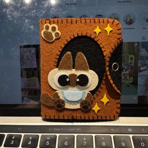 Puede incluir: Funda de teléfono de fieltro hecha a mano en tonos marrón, negro y crema, con un diseño caprichoso de gato. La funda tiene una huella de pata y detalles de estrellas, con cierre de botón a presión. El gato tiene ojos grandes y un babero.