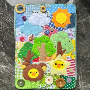 May include: A colorful, patchwork iPad case featuring a whimsical landscape design. The case includes felt appliqués of a sun, clouds, trees, and animal faces, with various buttons and fabric patterns. The case is designed to fit an iPad.