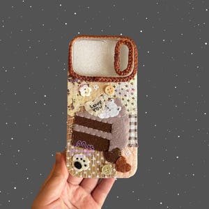 May include: A brown and beige phone case with a felt cake design. The case features a slice of chocolate cake with white frosting, buttons, and the text "Love You". The case has a woven brown border.