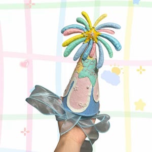 Puede incluir: Un gorro de fiesta hecho a mano en colores pastel. El gorro tiene una base azul, detalles de fieltro rosa y blanco, y una parte superior multicolor en forma de estrella. Una cinta azul transparente está unida a la base. El gorro tiene la inscripción "Happy Birthday".