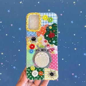 May include: A patchwork phone case with a grey Totoro character, colourful fabric squares, and decorative buttons. The case has a clear phone camera opening and a yellow braided border. The design includes a felt tree and a red apple.