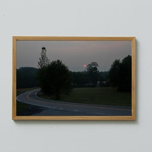 May include: A framed landscape photograph featuring a winding road through a rural area. The image captures a dusky sky with a red sun, trees, and a grassy field. The frame is made of light-colored wood.