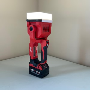 May include: A red and black cordless work light with a white light panel and a Milwaukee logo. The light is on a white surface.