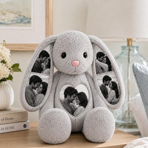 May include: A plush gray rabbit stuffed animal featuring a pink nose and black eyes. The rabbit has three heart-shaped photo inserts: one on each long ear and one on the center of its chest, each displaying a black and white photograph of a couple.