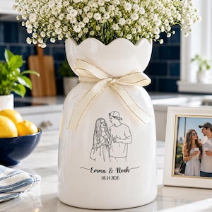 May include: A white ceramic vase with a scalloped rim and a cream-coloured ribbon tied around the neck. The vase features a black line drawing of a couple, the text "Emma & Noah", and the date "06.14.2026" printed on the front surface.