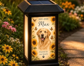 May include: A black solar-powered garden light featuring a memorial plaque with a golden retriever portrait, sunflowers, and butterflies. The text reads 2011-2026, Max, and Forever in our hearts, accompanied by a small paw print icon at the bottom.