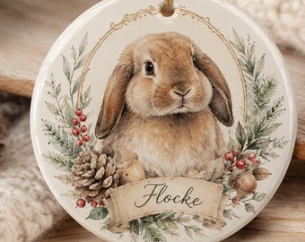 May include: A round white ceramic ornament featuring a brown lop-eared rabbit illustration centered within a decorative wreath of pine branches, red berries, and pinecones. A beige banner at the bottom displays the name Flocke in black script.