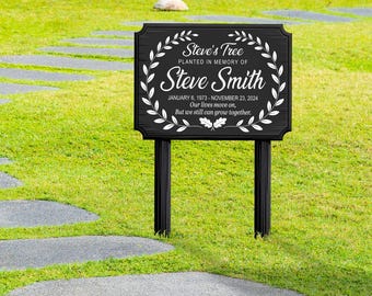 May include: A black memorial sign with the text "Steve's Tree" and "Steve Smith" in white script, surrounded by a white wreath. The sign includes dates and a message, mounted on black posts in a grassy yard.