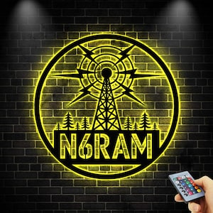 May include: A black metal wall art piece with a yellow neon glow. The design features a radio tower, lightning bolts, and the text "N6RAM" inside a circular frame. The background is a black brick wall.